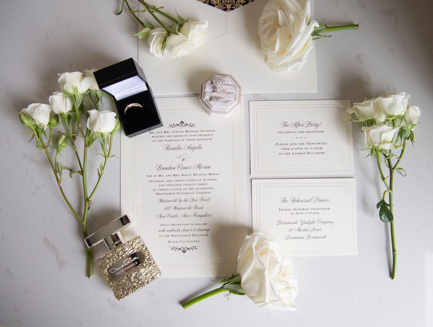 Elegant wedding invitation suite with white roses, spray roses, and perfume bottle arranged on white marble surface.