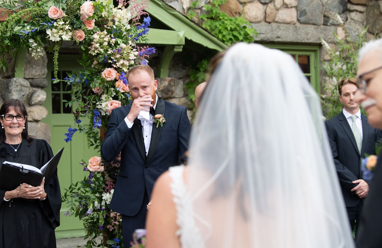 Wedding ceremony setup outdoors with floral arch and stone wall backdrop.