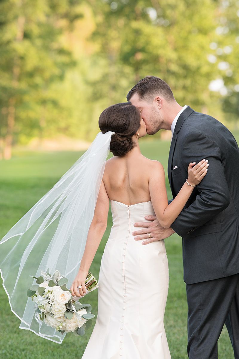 Elegant wedding couple embraces outdoors with flowing veil and white bouquet against green garden backdrop.