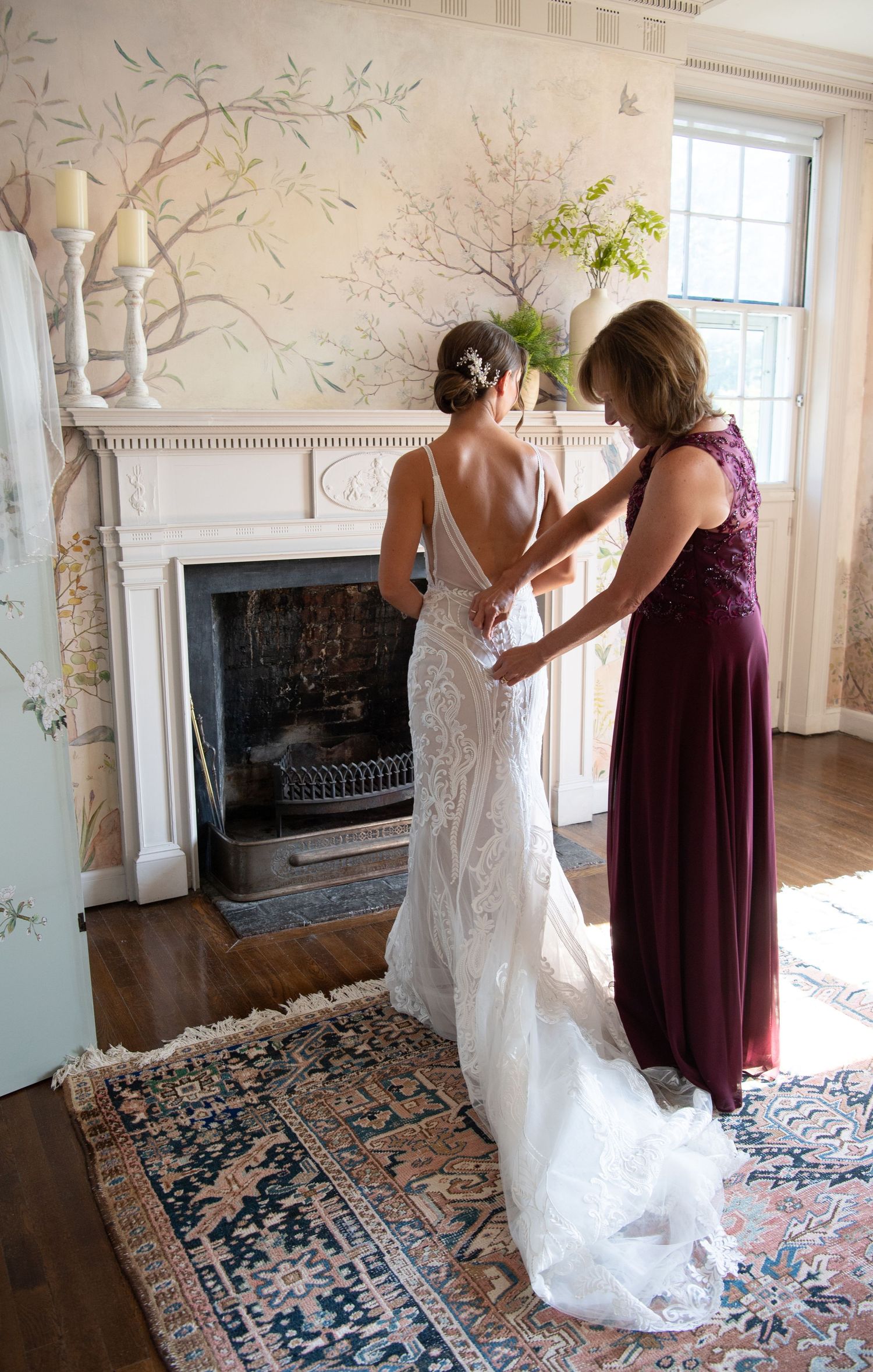 Wedding preparations in an elegant room with white walls adorned with delicate tree branch artwork and a vintage fireplace.
