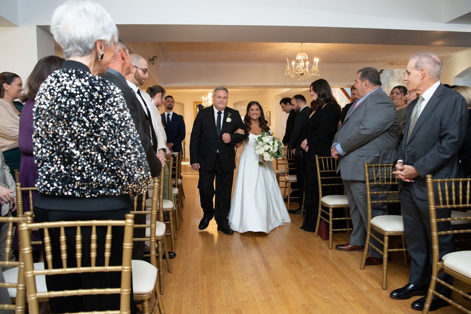 Wedding guests gather in an elegant reception hall with white decor and chandeliers for a celebration.