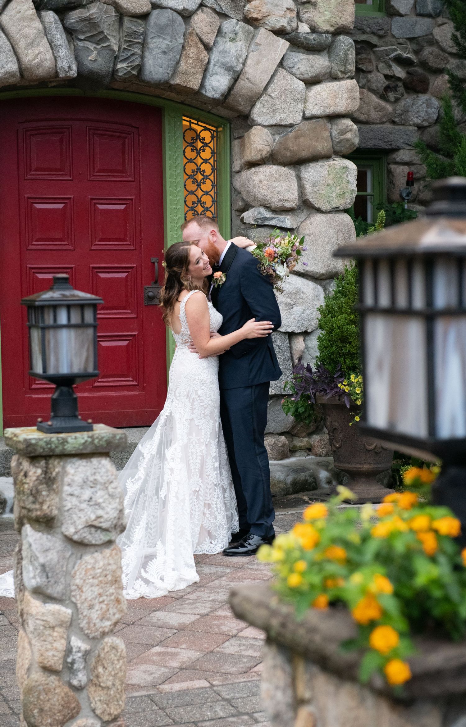 A romantic moment captured in front of a red door and stone wall with yellow flowers in the foreground.
