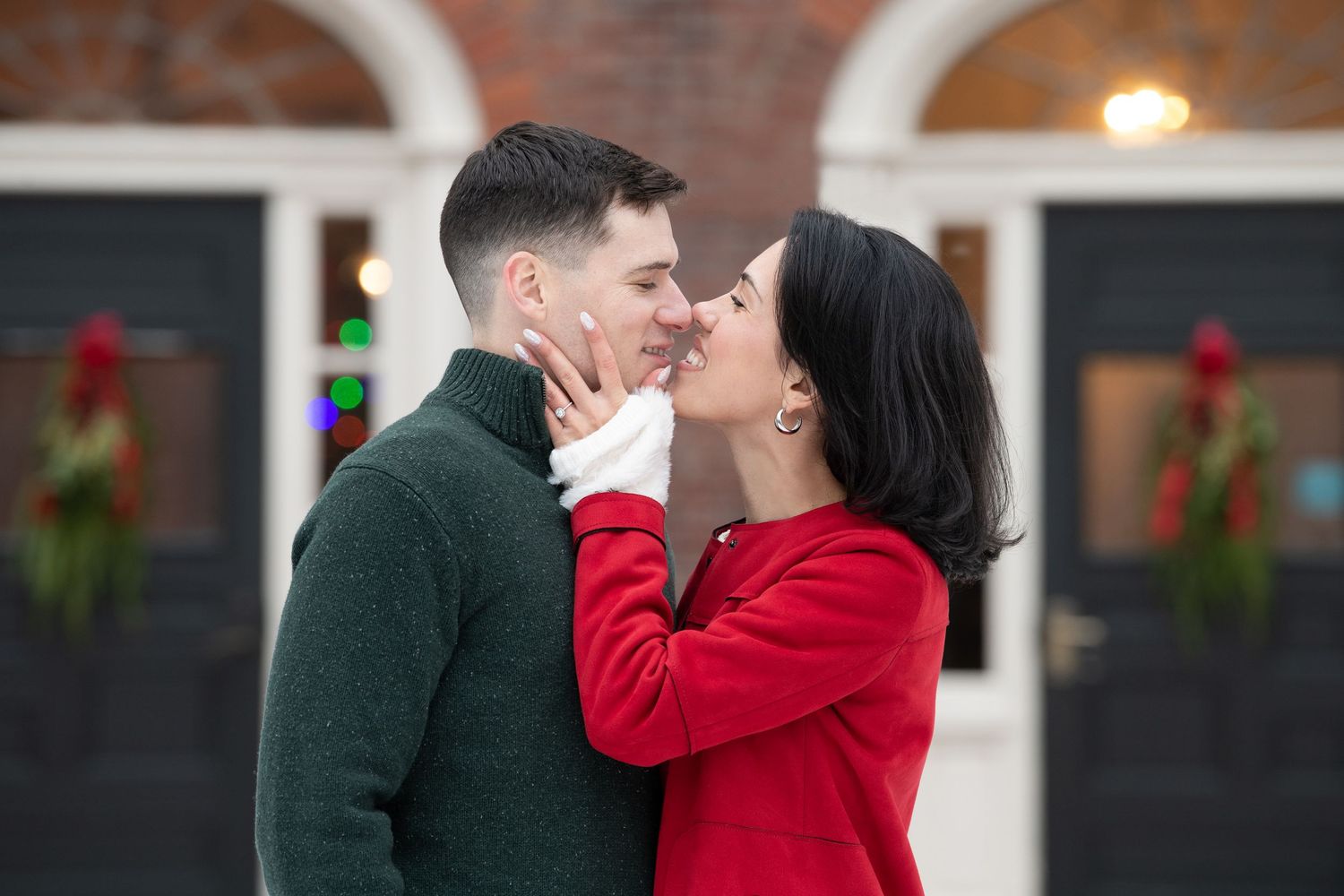 A romantic couple in winter attire share a joyful moment outside a brick building with holiday decorations.