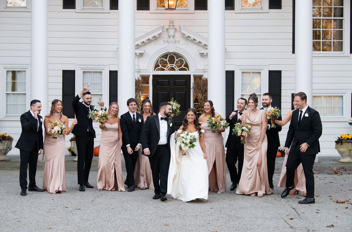 Wedding party in formal attire poses together in front of a white church with elegant columns and windows.
