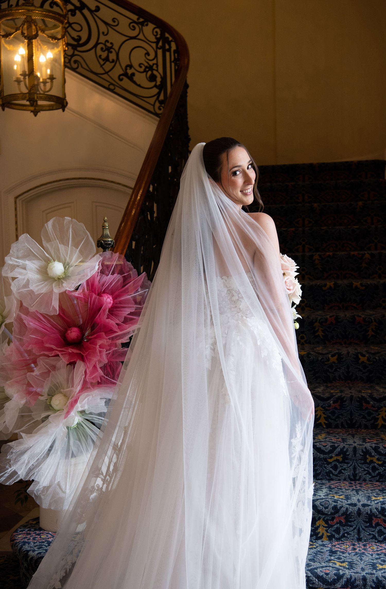 Bride in a white lace gown and long veil stands on an elegant staircase holding a bouquet, smiling gracefully.