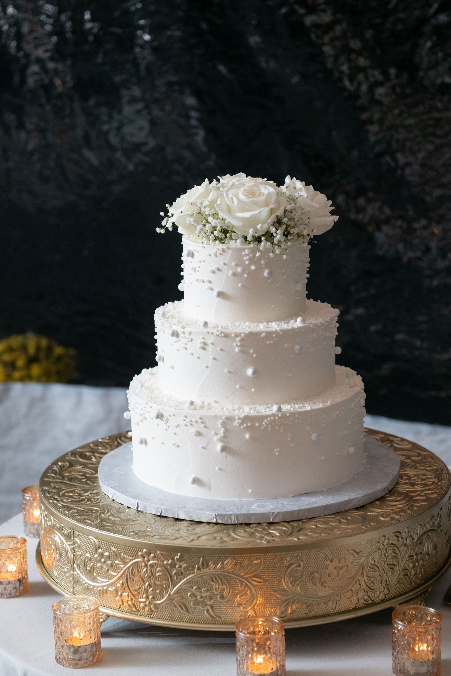 An elegant three-tier white wedding cake decorated with roses and baby's breath sits on an ornate gold stand with glowing votives.