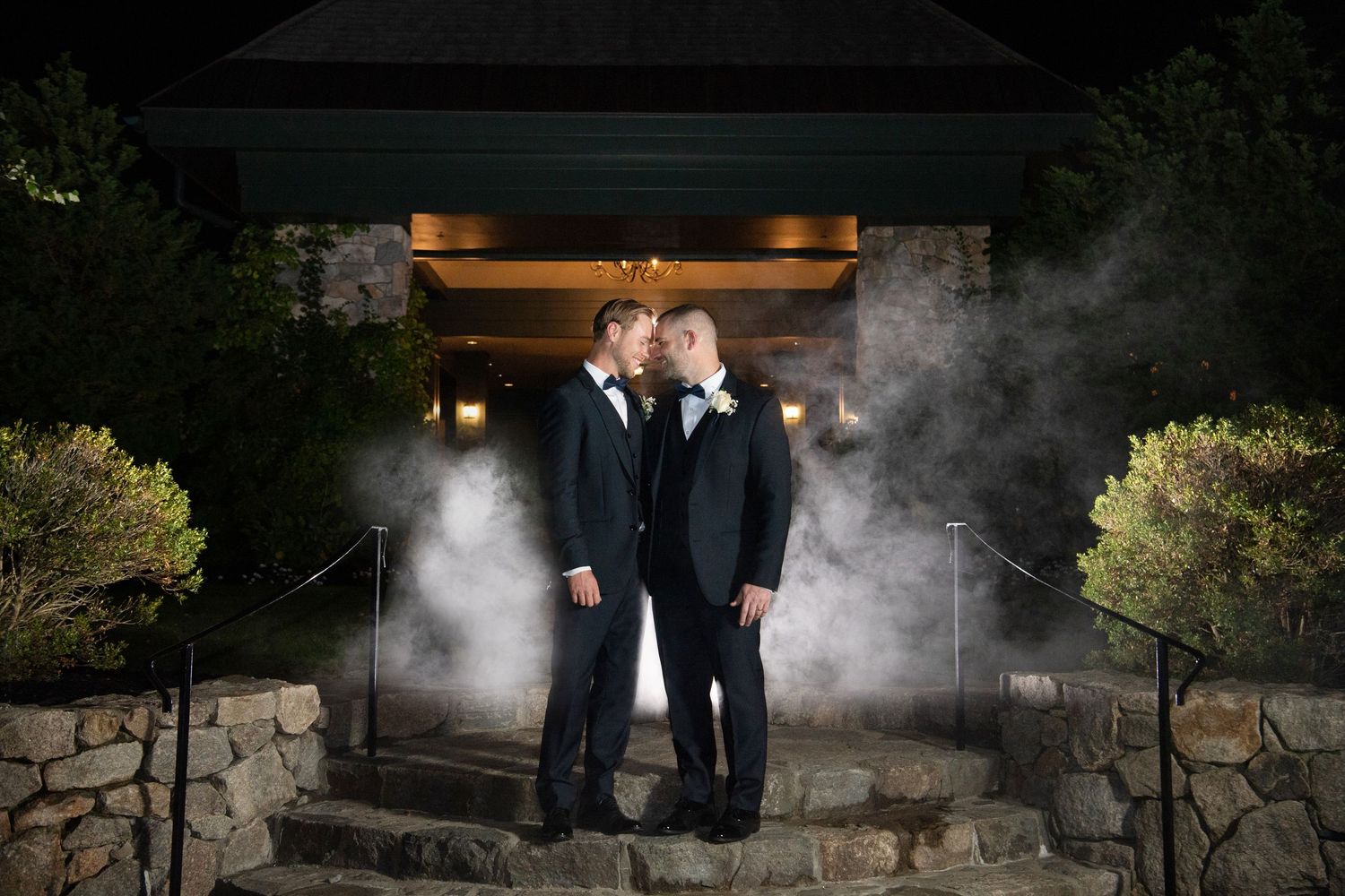 Two formal-dressed figures standing near a misty garden path with architectural elements at night.