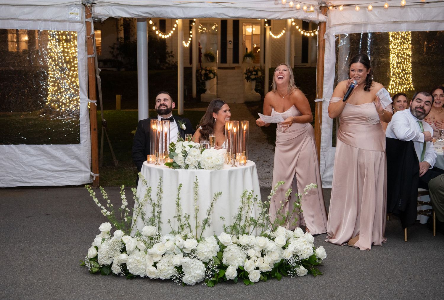 Wedding reception decor features elegant white floral centerpieces and soft pink bridesmaid dresses at an indoor venue.