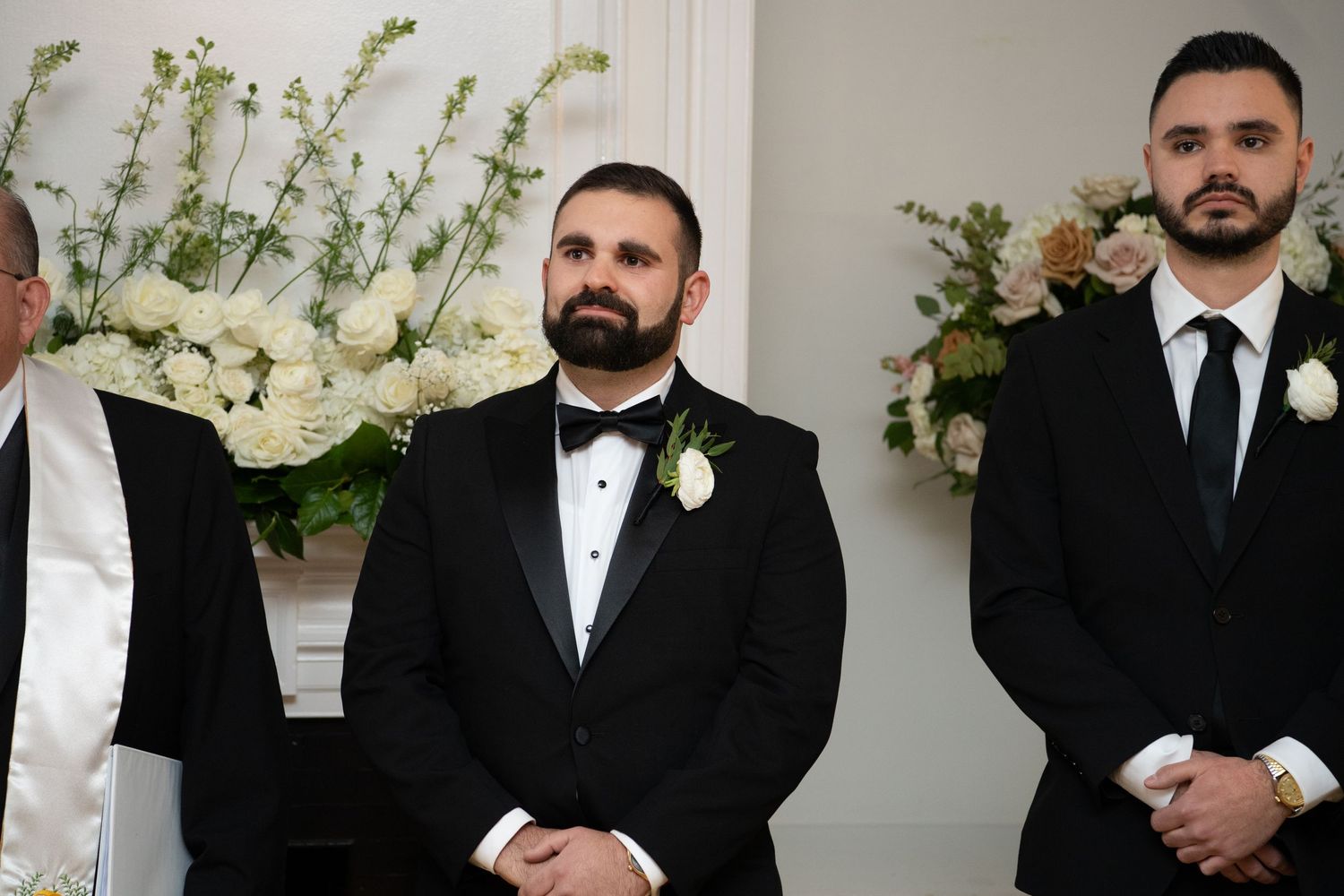 A wedding party in black suits and white shirts poses in a line against a white wall with floral decorations.