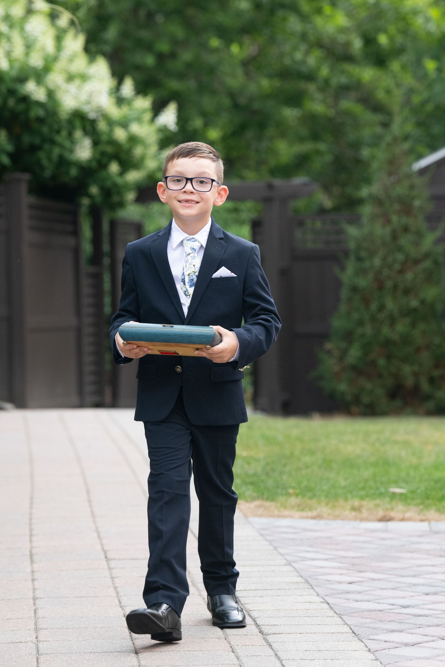 A young ring bearer in a formal black suit and glasses carries a pillow while walking down a garden path.