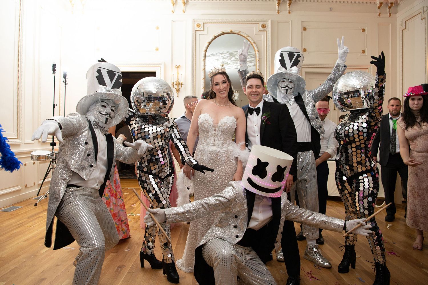 Couple poses with Marshmello DJ and silver robot dancers at elegant wedding reception party.