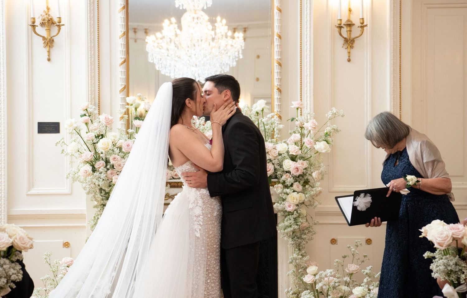 Bride and groom share first kiss at elegant wedding ceremony with white floral arrangements and chandelier backdrop.