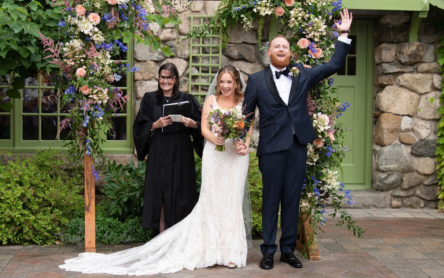 Wedding party stands in front of stone wall with pink climbing roses and green door at rustic garden venue.