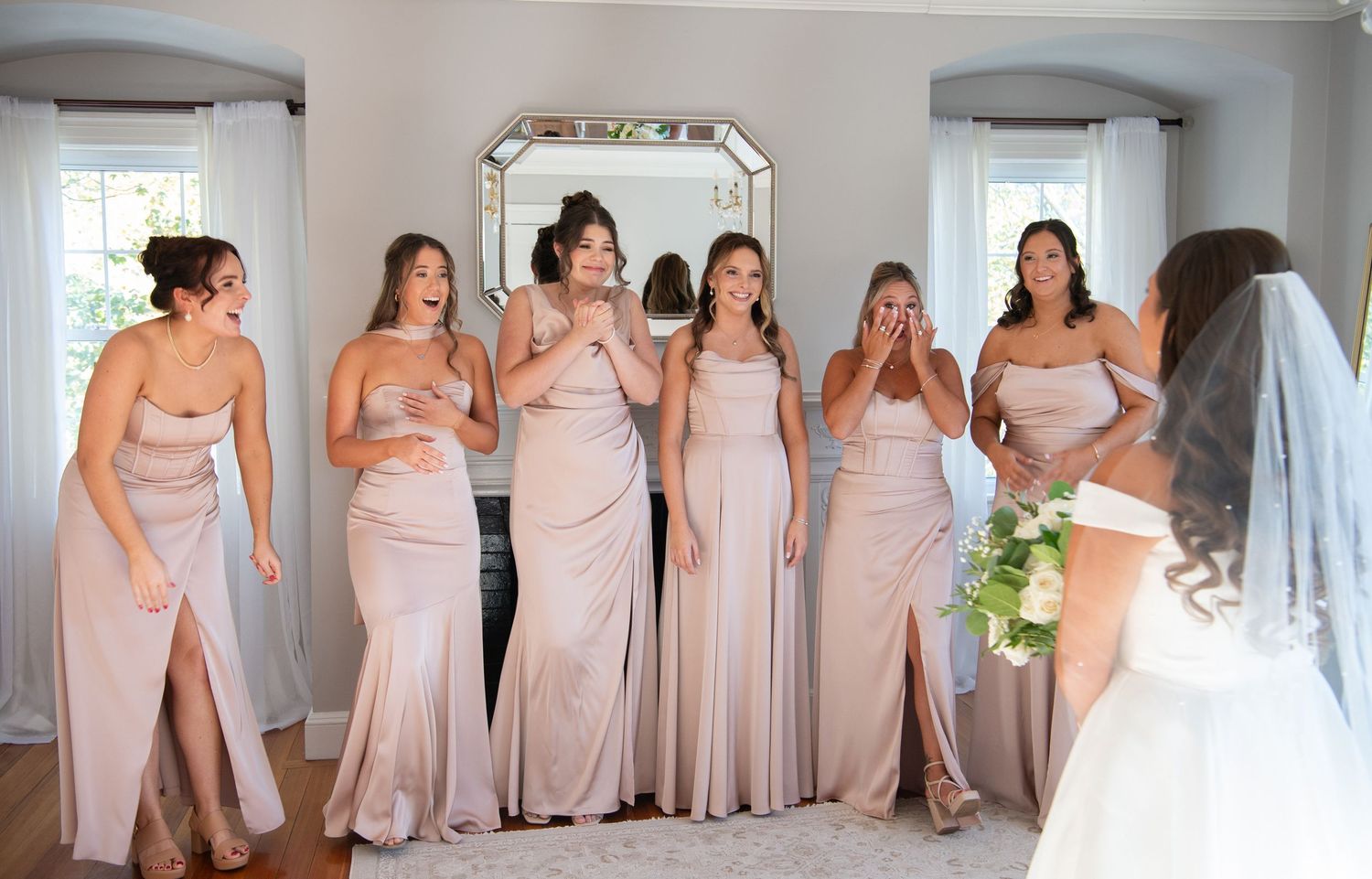Bridesmaids in blush pink dresses pose together in a line against a white wall during a wedding celebration.