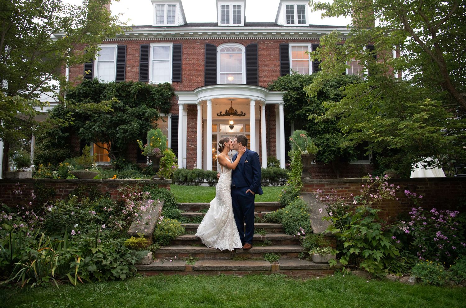 A classic brick mansion with white columns and dormer windows sits behind a manicured garden path.