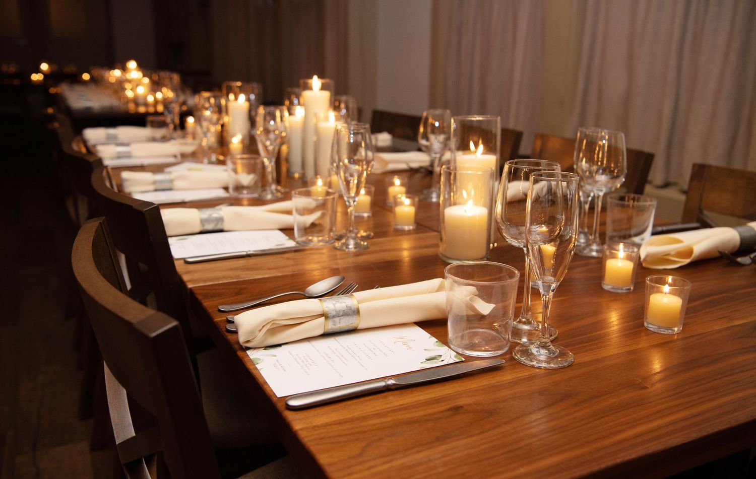 Elegant dinner table setting with glowing candles and wine glasses on dark wooden surface in dim lighting.