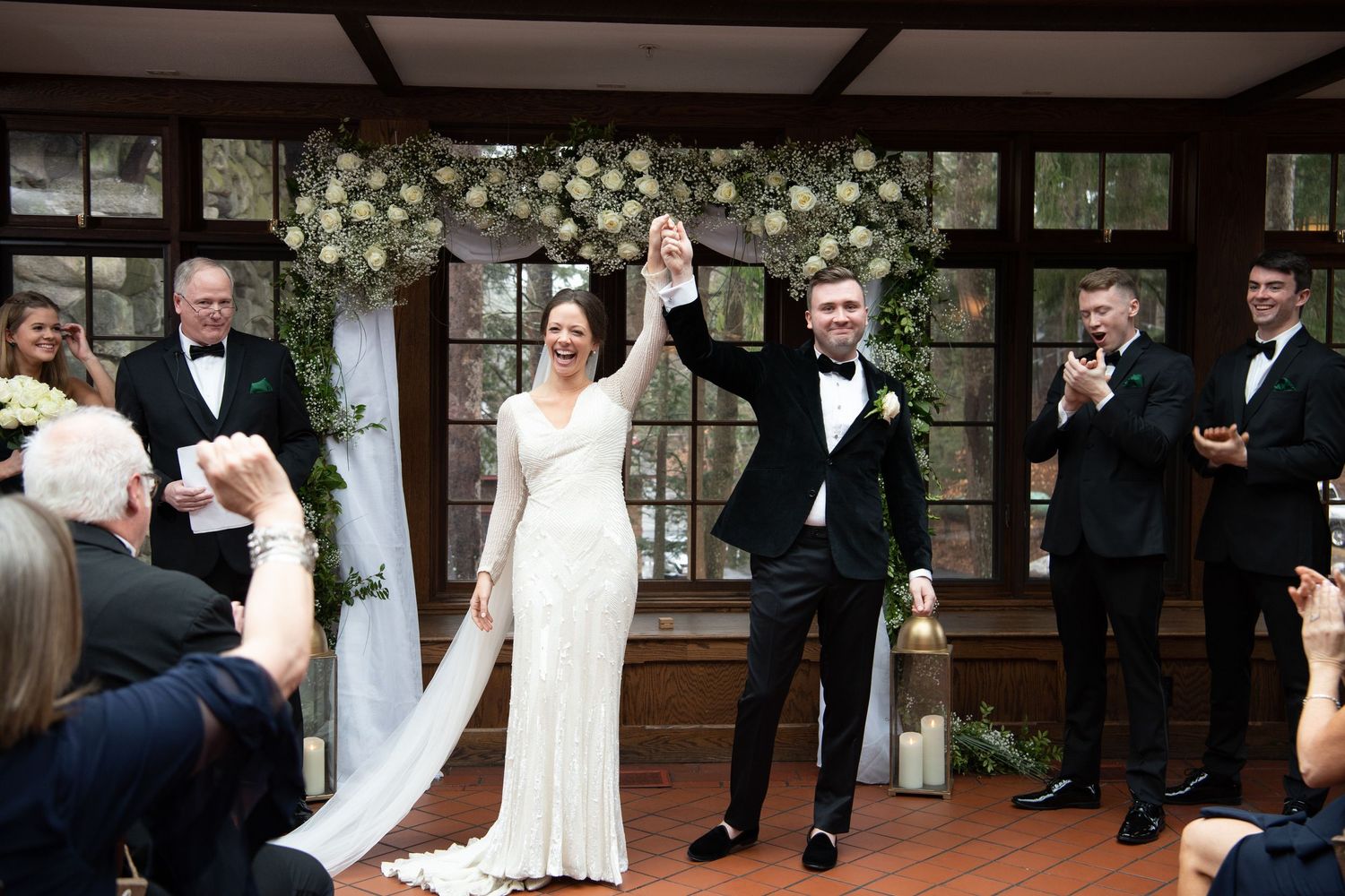 Wedding party members talk and laugh together on a wood deck patio during an outdoor celebration.