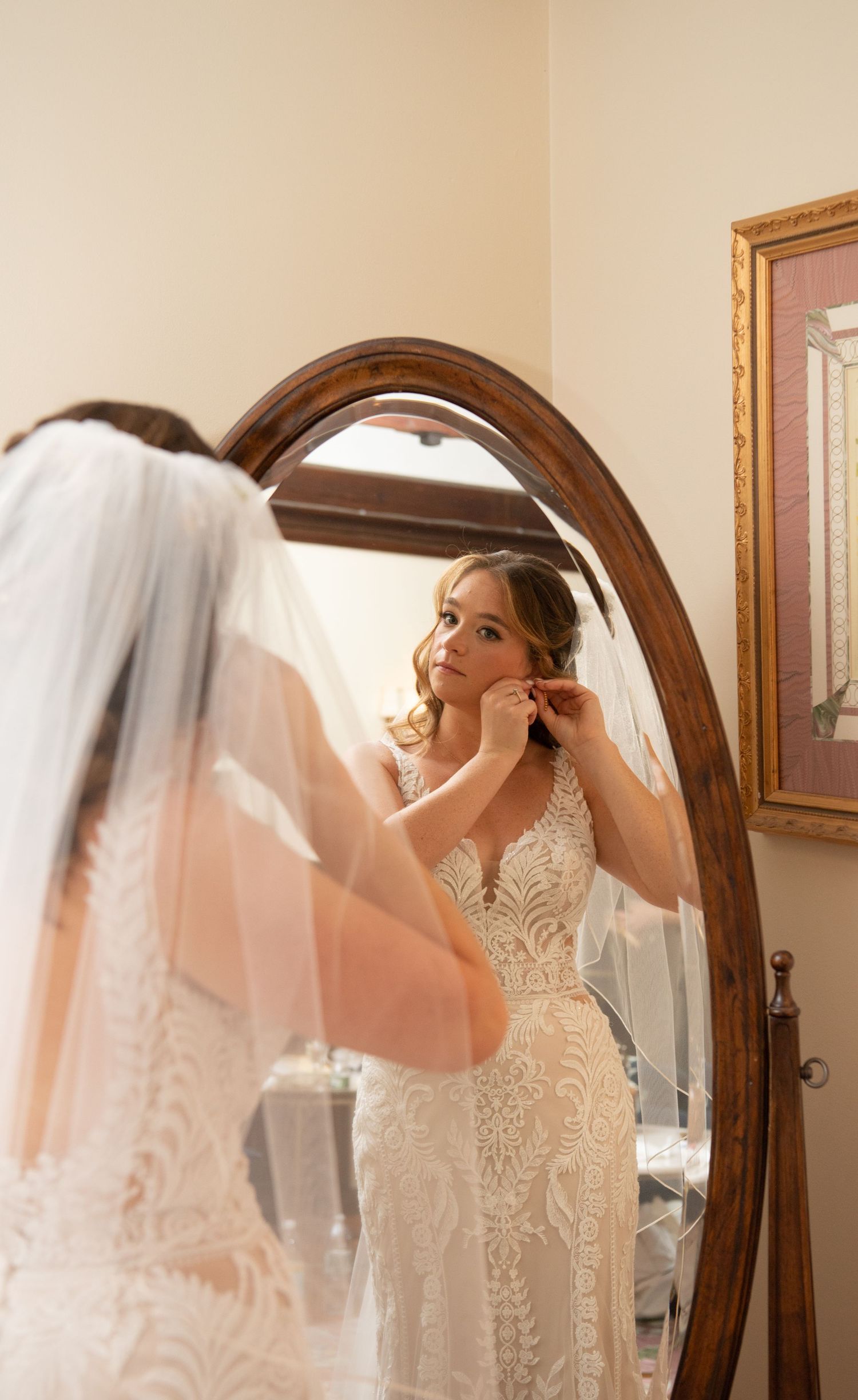 Bride in lace wedding dress putting on earrings while looking in an ornate oval mirror.