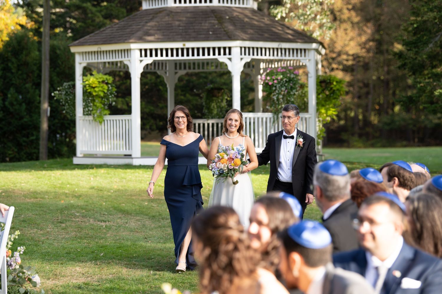 Wedding guests gather outdoors near a white gazebo on a sunny afternoon.