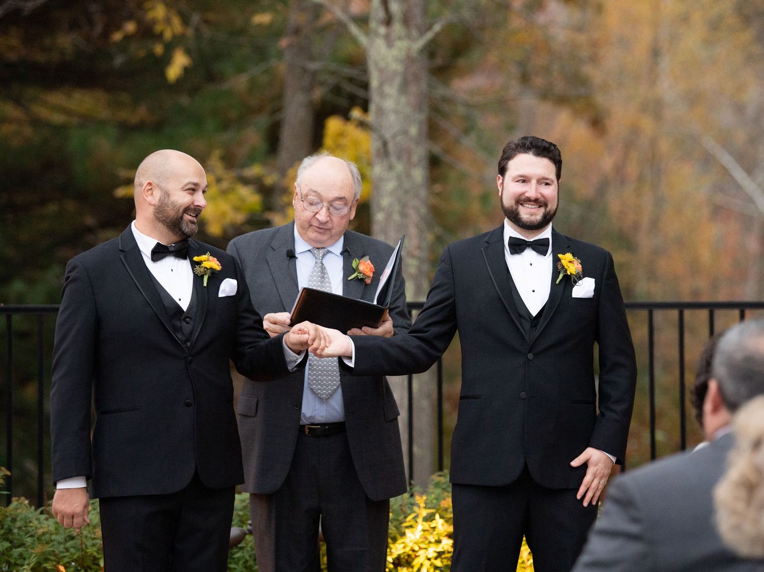 A group in formal black tuxedos and suits share a lighthearted moment during an outdoor autumn wedding.