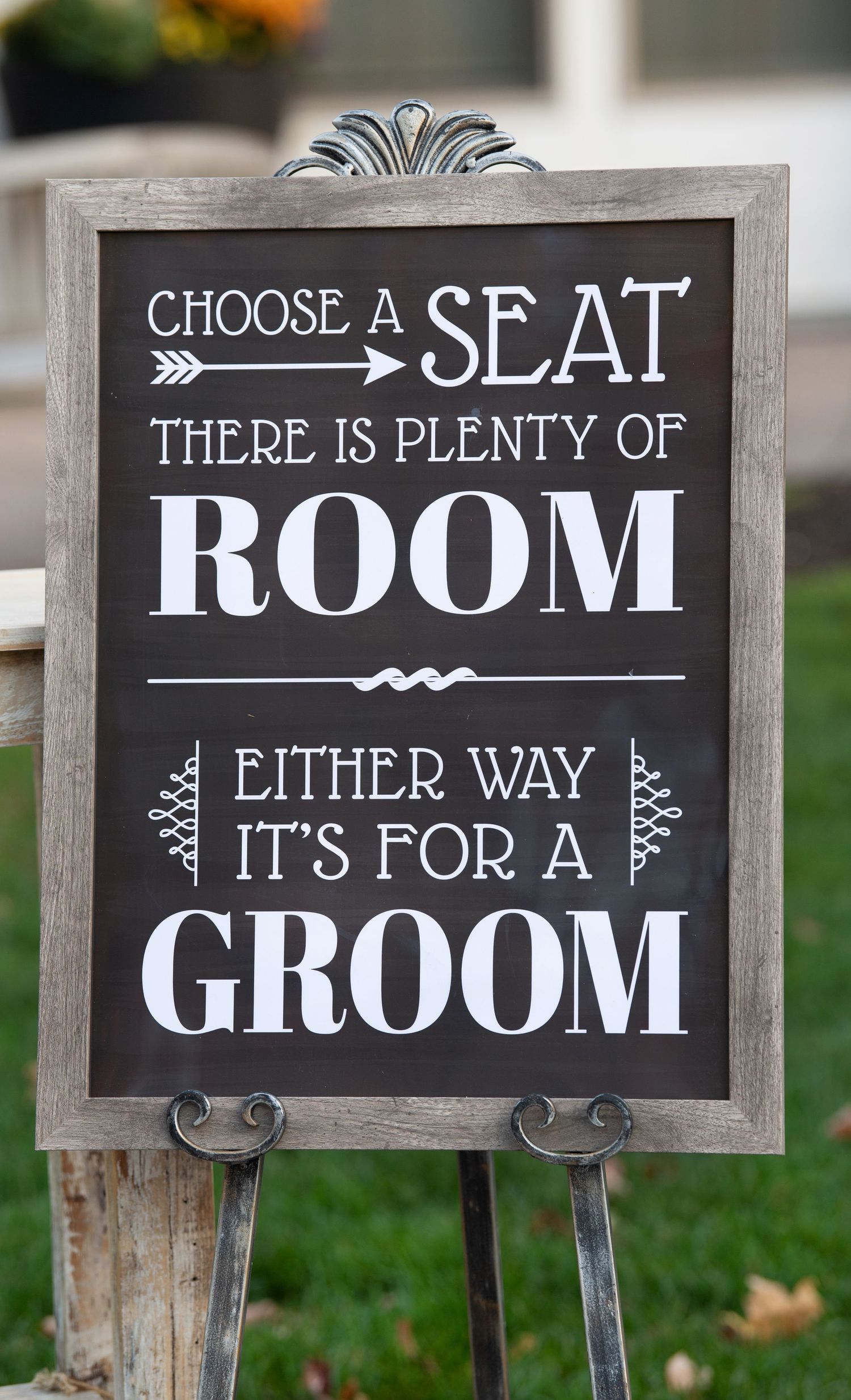 A rustic framed chalkboard wedding sign with text reading 'Choose a seat there is plenty of room either way it's for a groom'.
