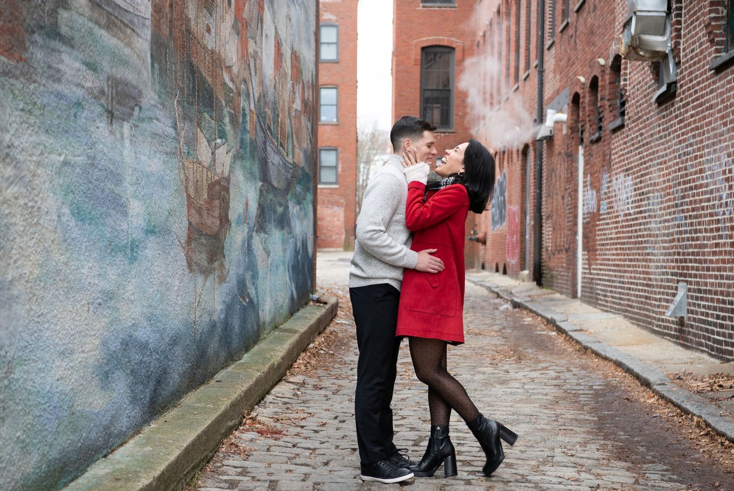 A couple embraces on a cobblestone alley between brick buildings in an urban setting.