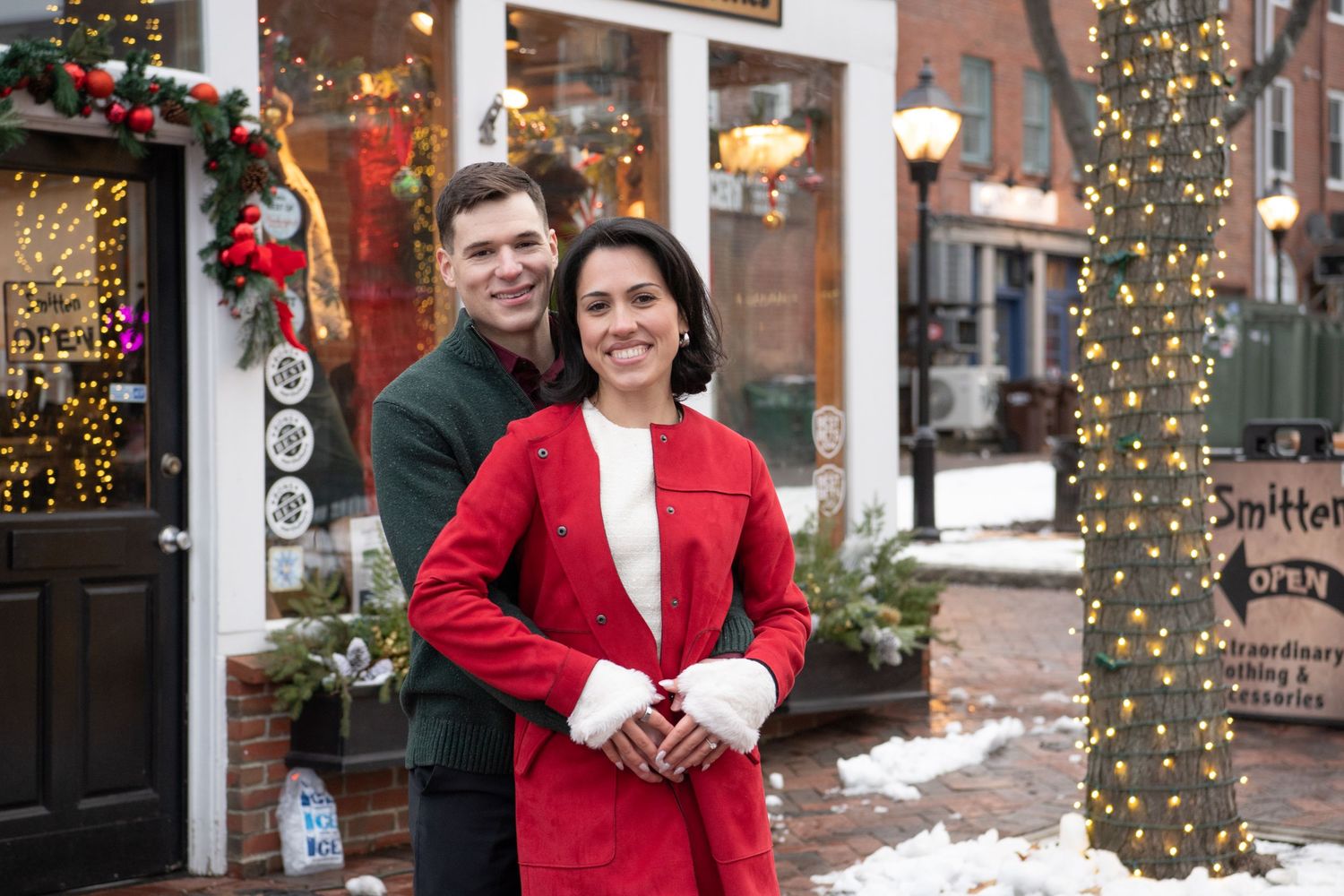 A couple in winter attire poses together on a snowy brick sidewalk with festive holiday decorations in the background.