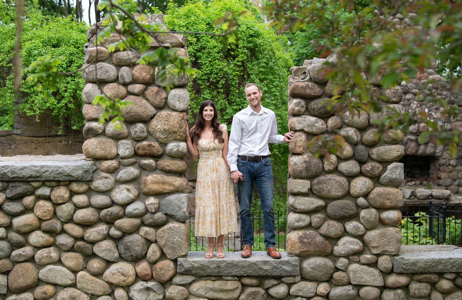 Couple standing beside a rustic stone wall in an outdoor garden setting during a summer engagement photoshoot.