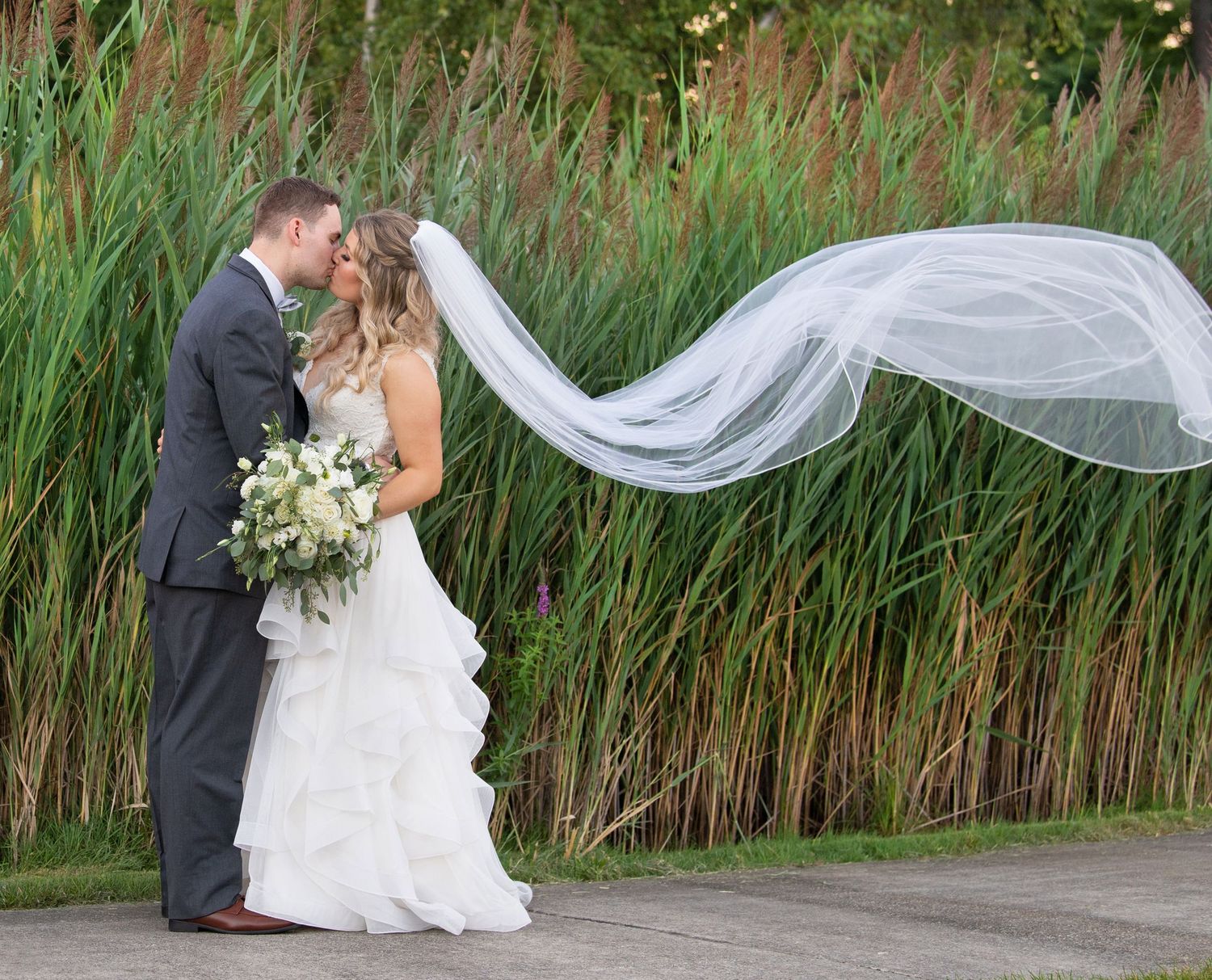A flowing white veil catches the breeze as newlyweds share a romantic moment against tall reeds and grass.