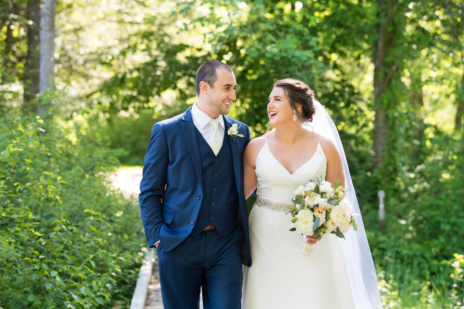 A happy couple in wedding attire walking through a lush green forest setting during a summer celebration.