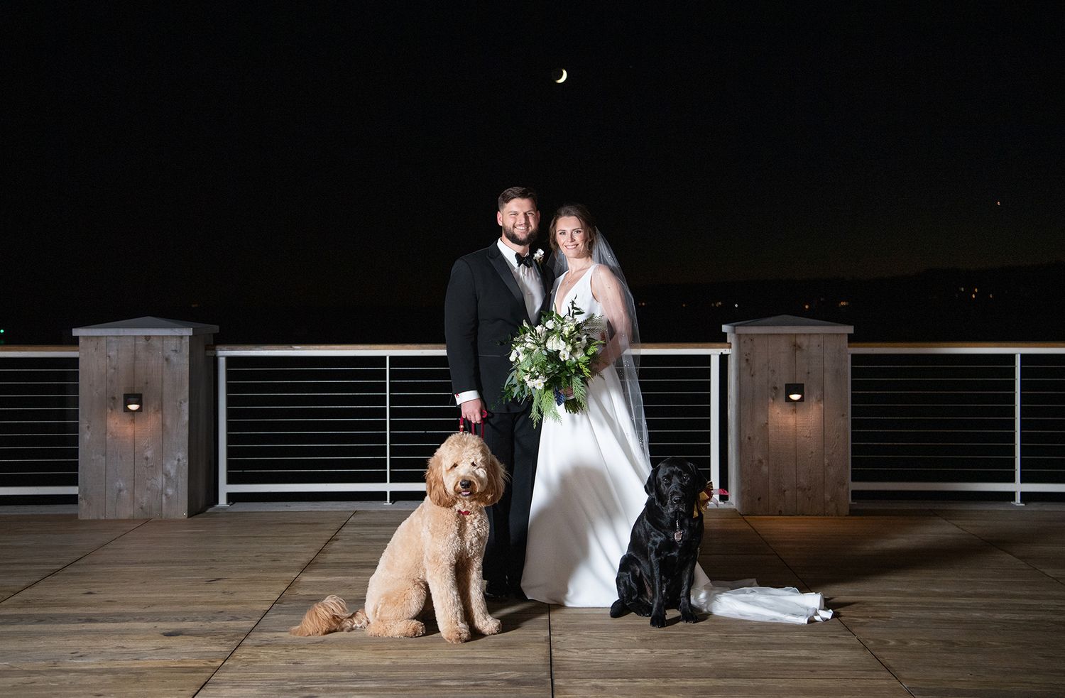A couple in wedding attire stands on a dimly lit wooden deck with wall sconces in the background.