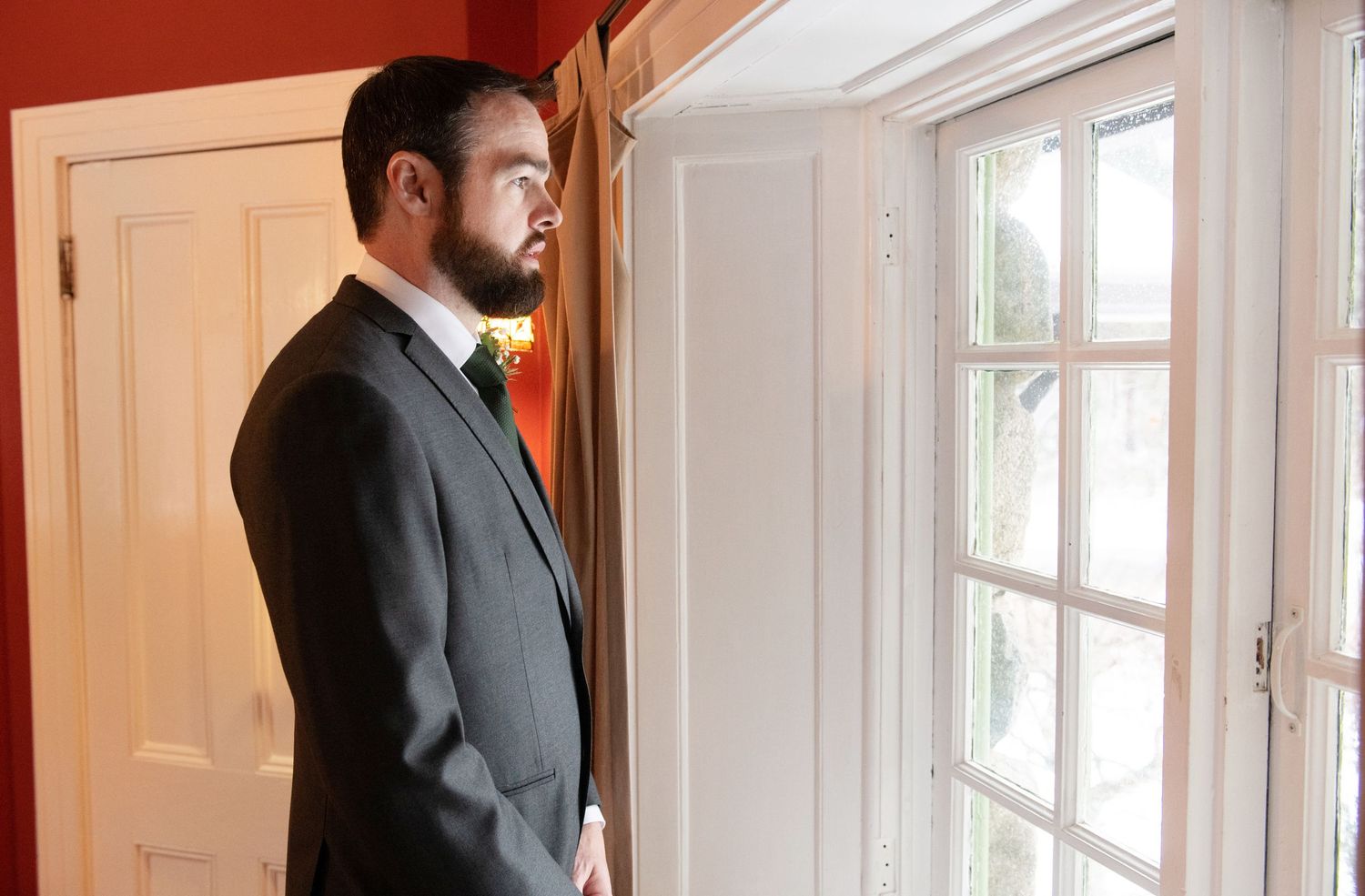 A bearded man in a dark suit stands contemplatively by a window with white panes.