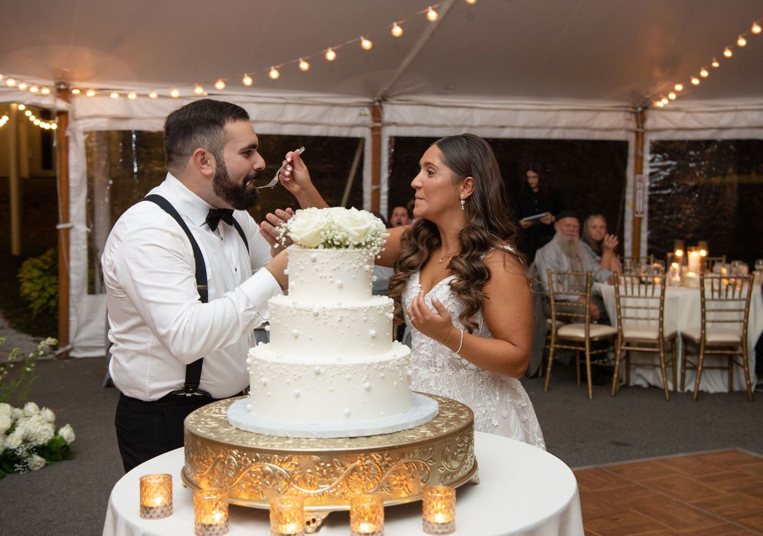 A wedding couple shares a romantic moment while cutting their white tiered wedding cake at an outdoor reception.