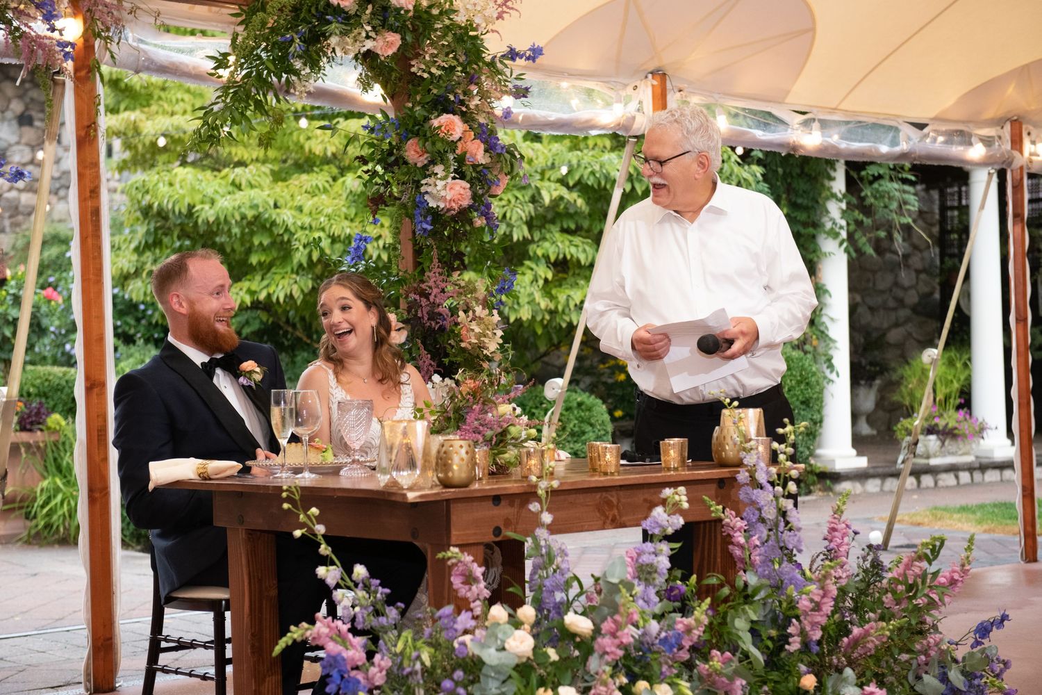 Outdoor wedding reception with wooden tables and hanging floral arrangements under tent canopy.