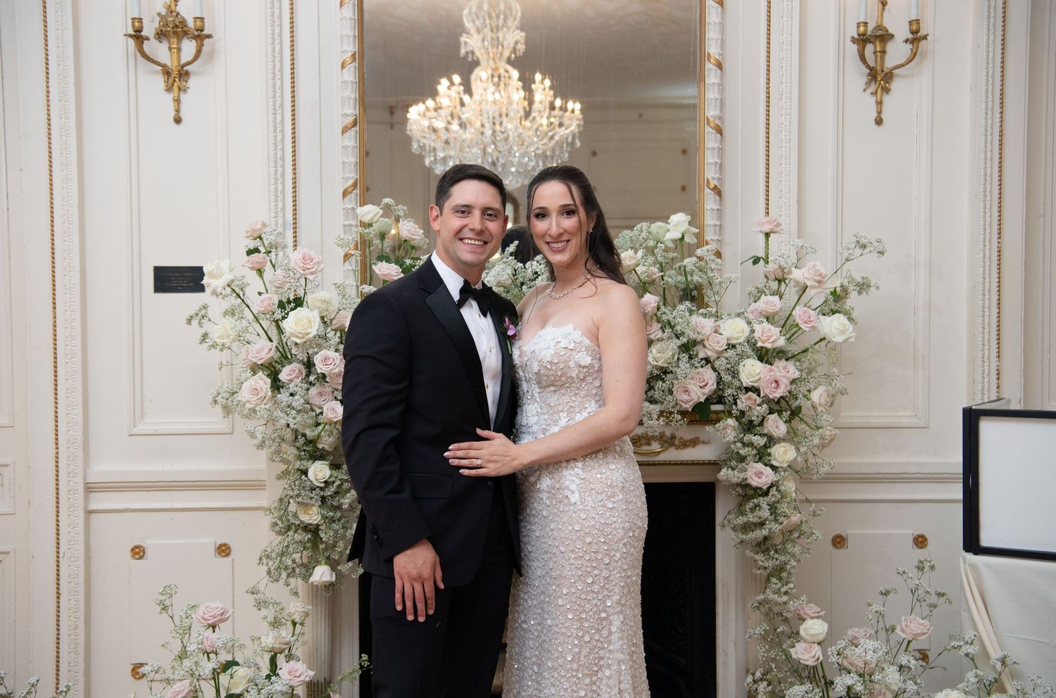 Elegant wedding couple posing before floral arch with pink roses, bride in beaded gown, groom in tuxedo, chandelier backdrop.