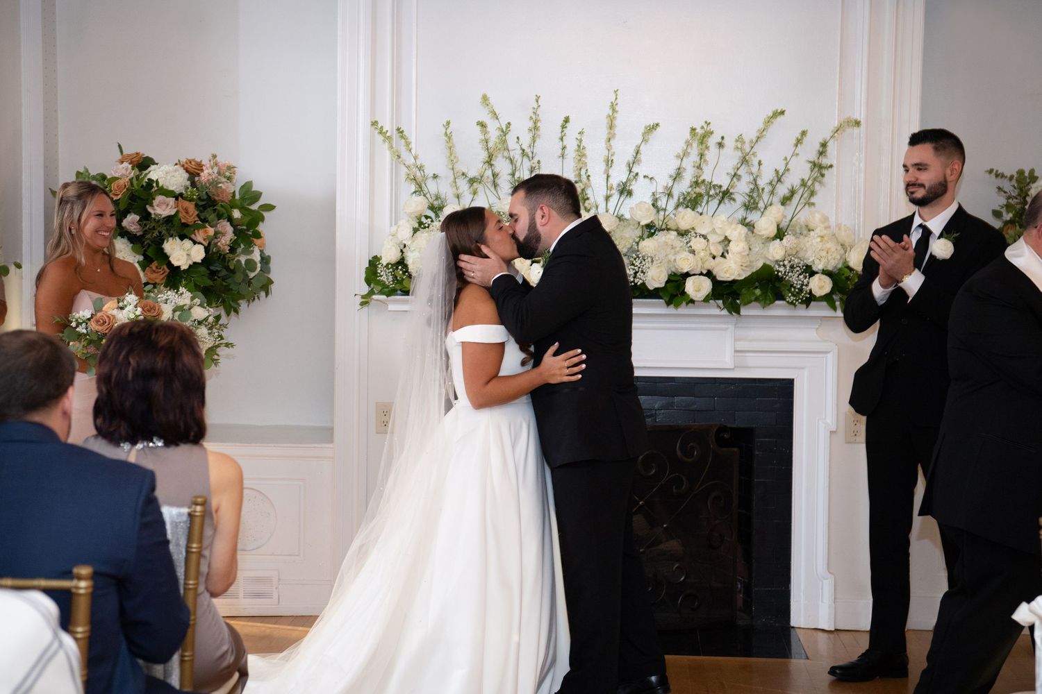 A couple shares their first kiss during a wedding ceremony in front of a white fireplace adorned with floral arrangements.