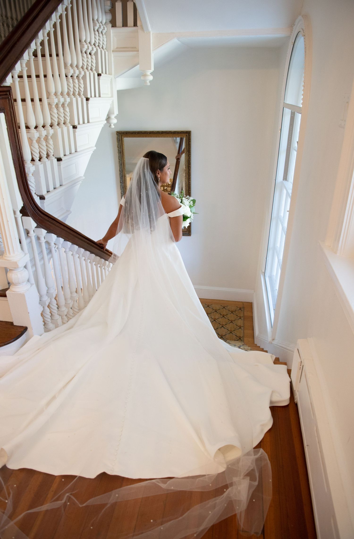 A flowing white wedding dress with a dramatic train displayed on wooden stairs in a bright interior setting.