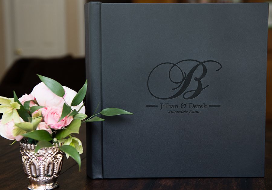 A black leather wedding album with monogram B next to a small vase of pink and white flowers.