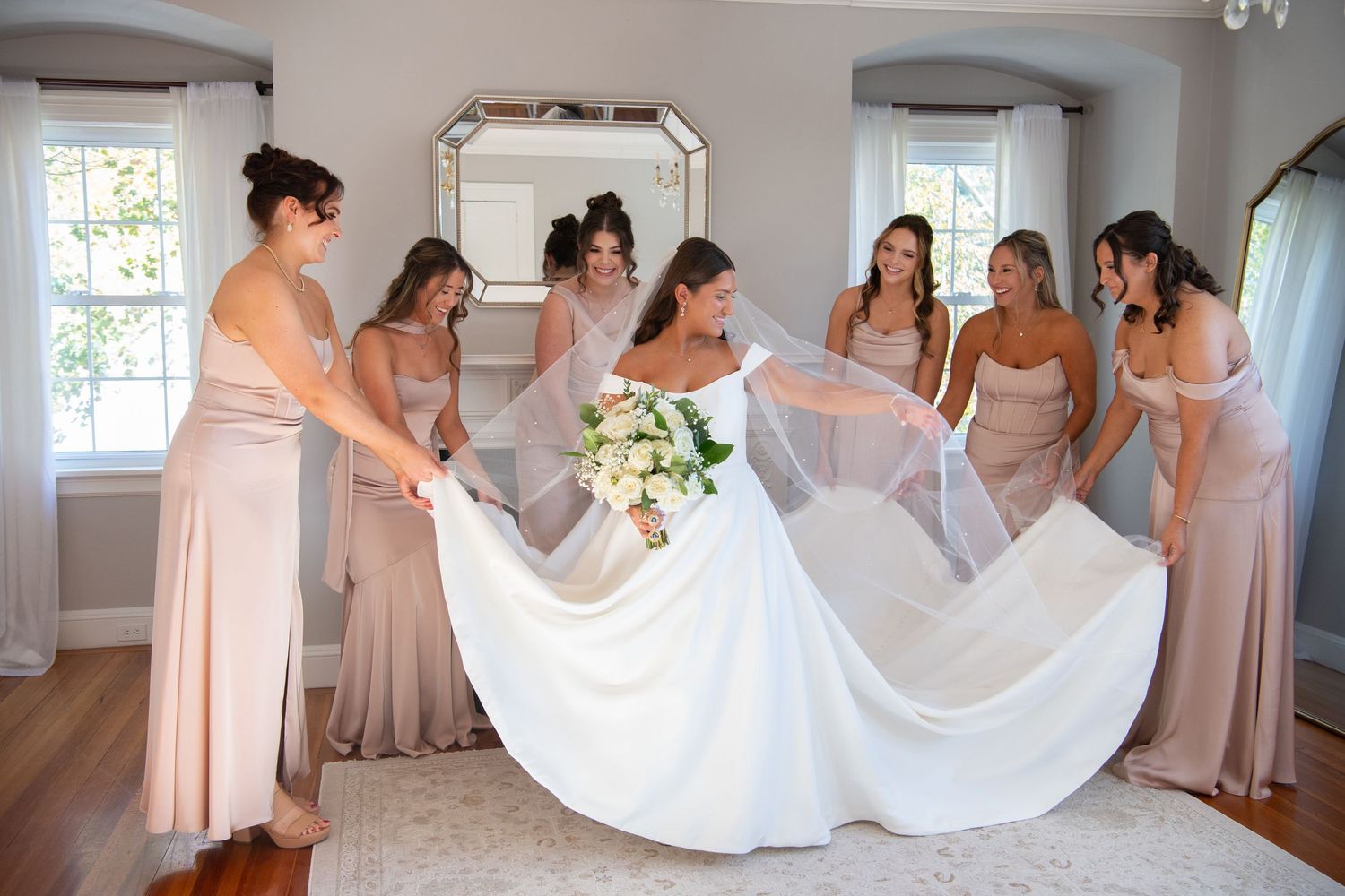 Bridesmaids in blush dresses help arrange a white wedding gown train in a sunlit room.