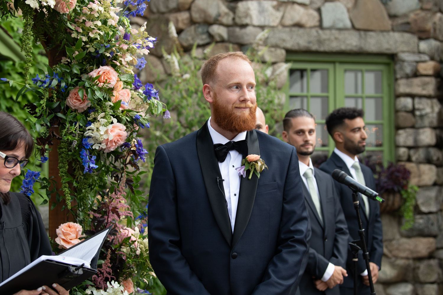 A floral wedding arch with pink and purple blooms stands against a rustic stone wall.