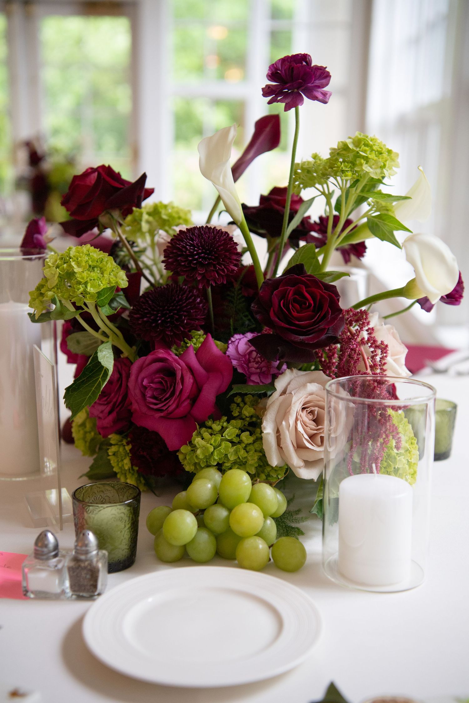 Elegant floral centerpiece with burgundy roses, calla lilies, green hydrangeas, and fresh grapes beside a white candle on a wedding table.