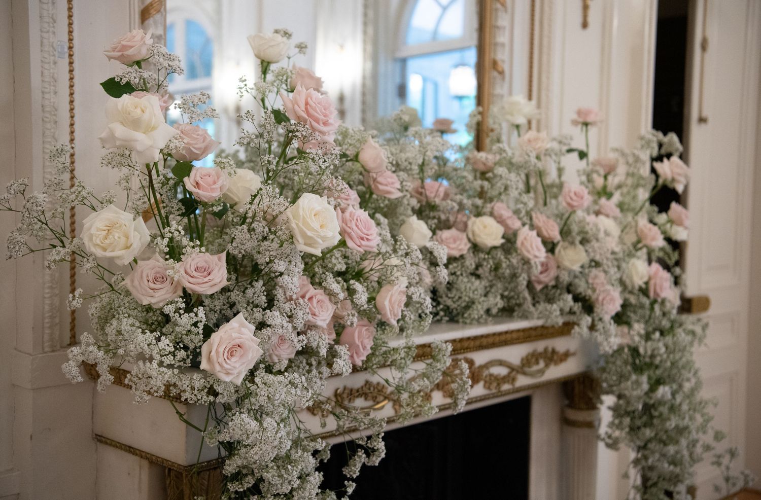Elegant fireplace mantel adorned with lush pink and white roses and baby's breath floral arrangement in a grand interior.