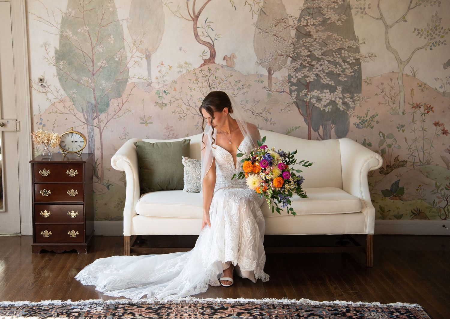 Bride in white lace gown holding colorful bouquet sits on ivory sofa before scenic woodland mural.