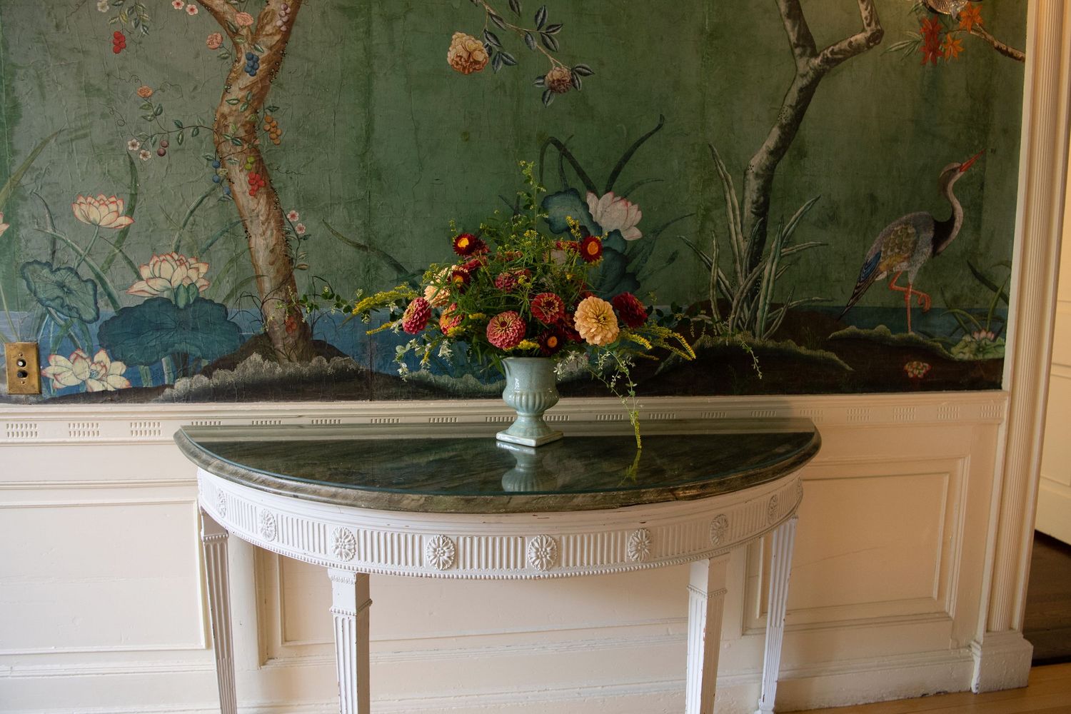 Elegant demilune console table with marble top holding a floral arrangement, set against a green chinoiserie wallpaper.