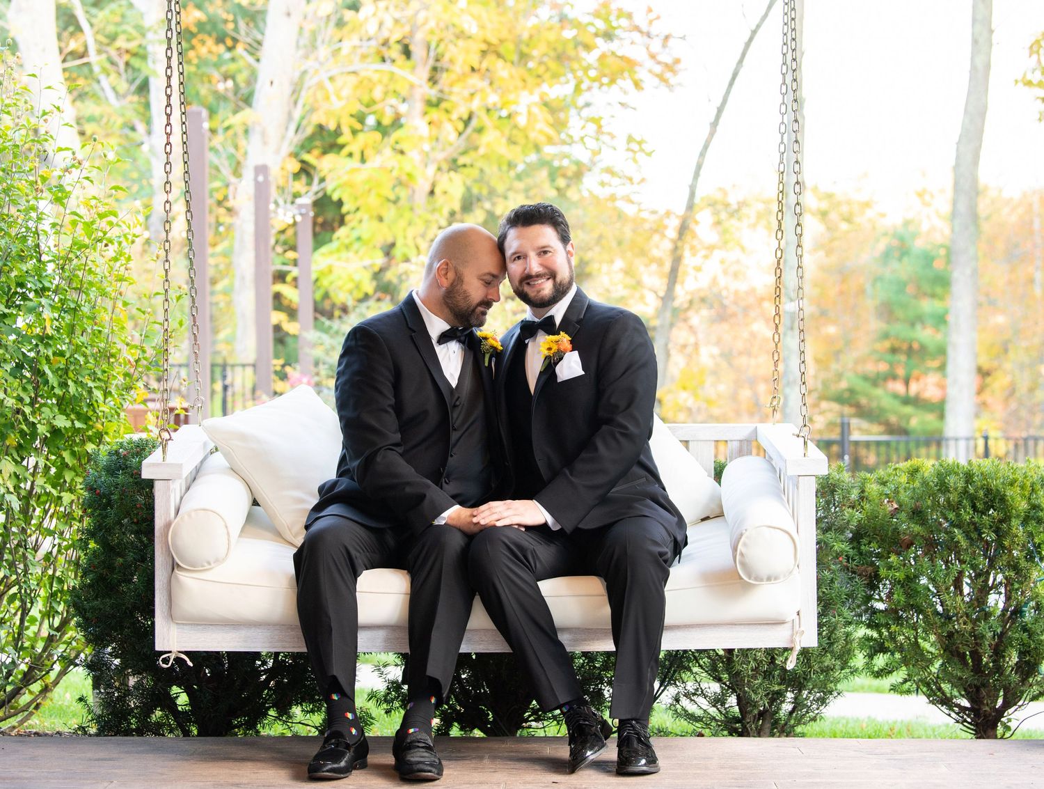 A happy couple in black tuxedos sit together on an elegant white porch swing surrounded by autumn foliage.