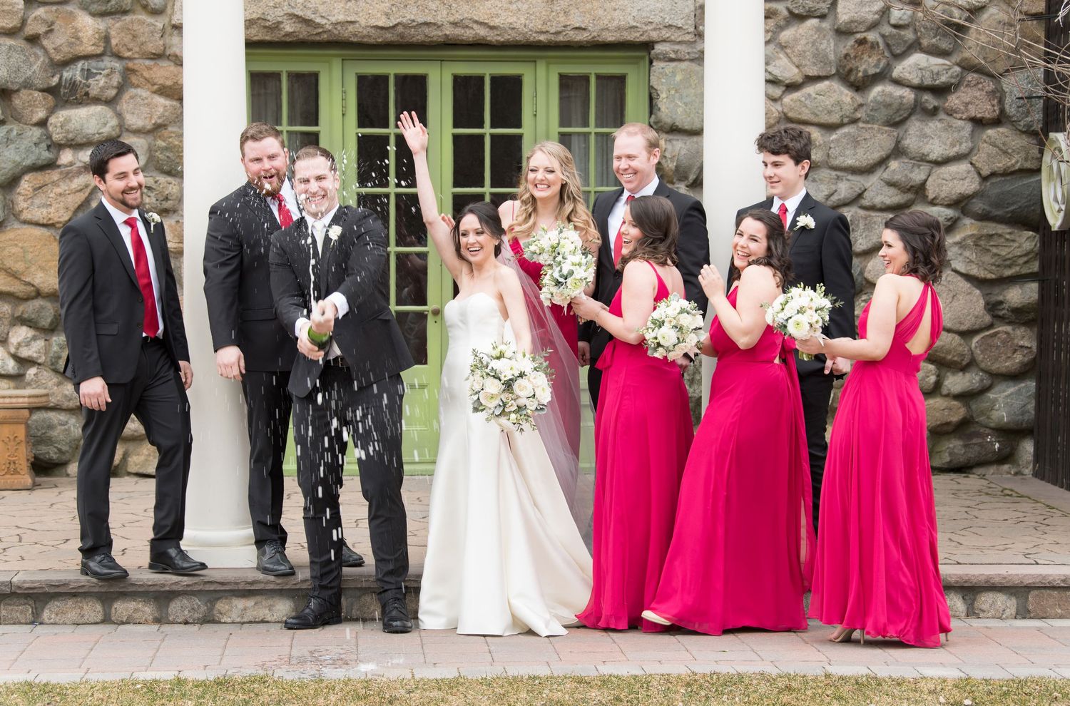 Wedding party lined up outside stone building wearing formal black suits and bright pink bridesmaid dresses.