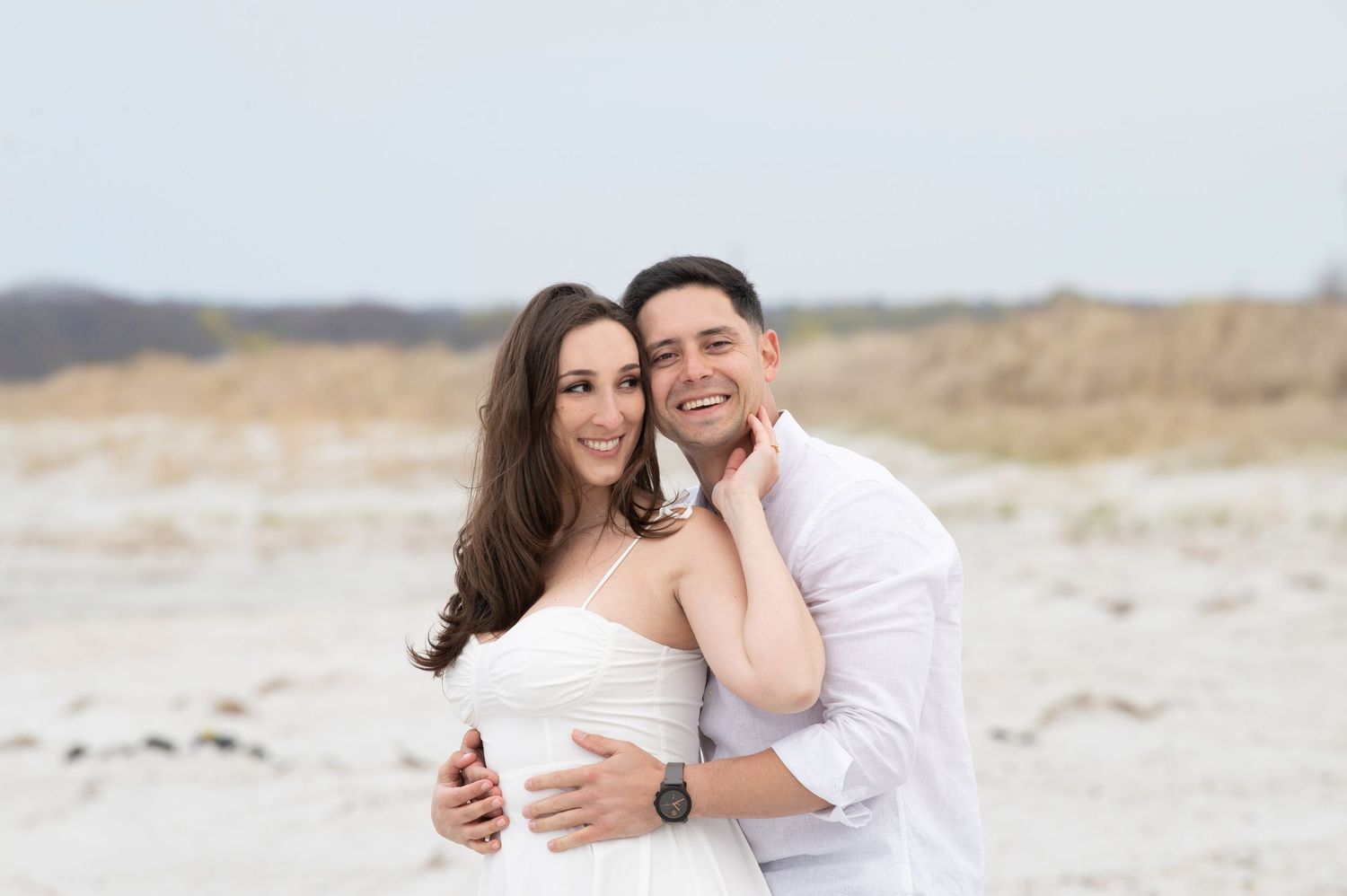 Couple in white outfits embracing during a romantic photoshoot on a sandy beach at golden hour.