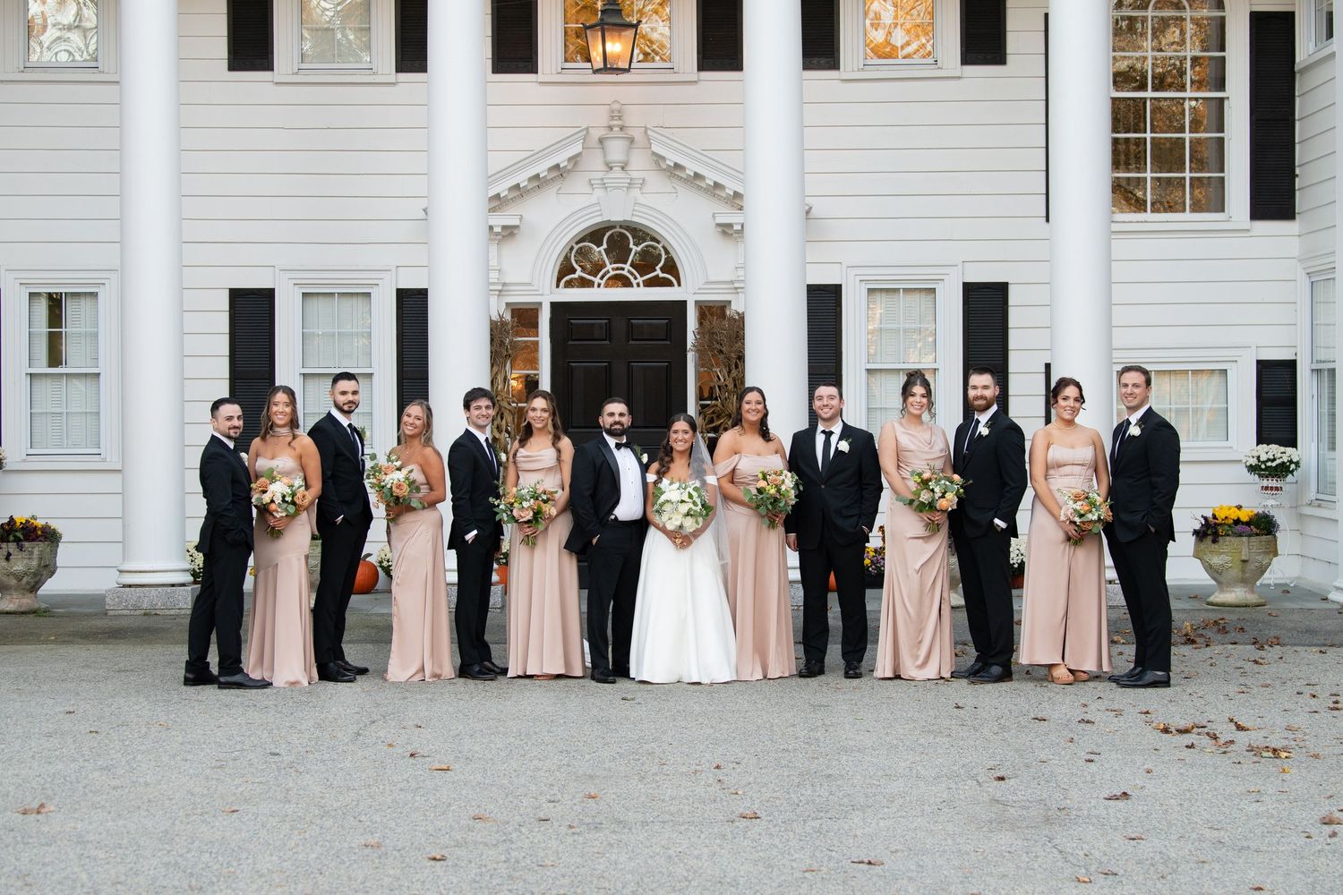Wedding party poses in front of elegant white mansion with black shutters and ornate doorway entrance.