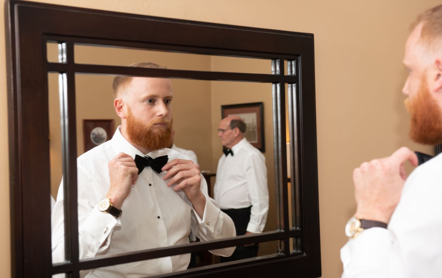 Groomsmen getting ready in formal black tuxedos and bowties while looking in a mirror.