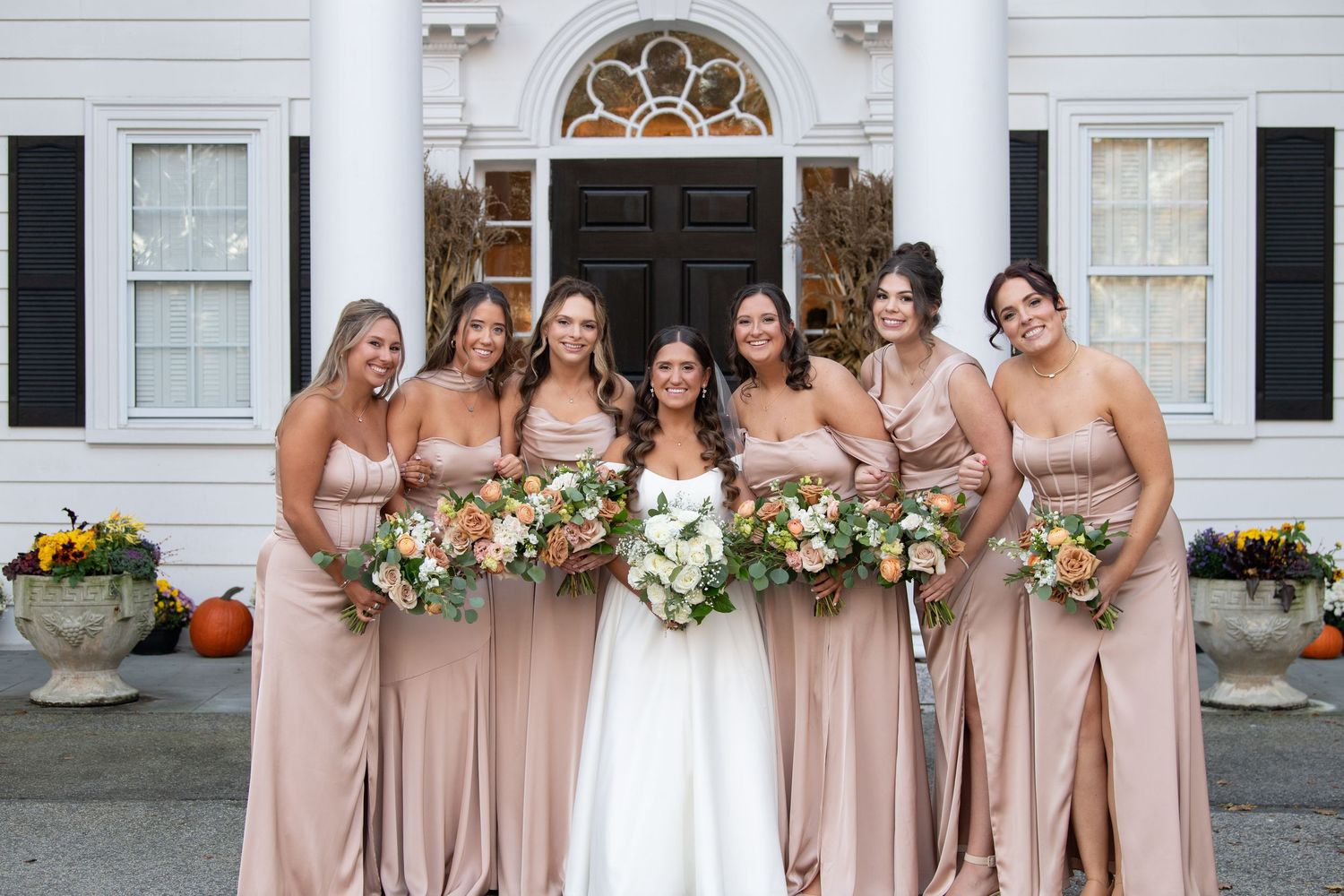 Bridesmaids in blush pink dresses pose with the bride on the porch of a classic white building.